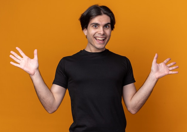 Überraschtes junges hübsches Kerl, das schwarzes T-Shirt trägt, das Hände verteilt auf orange Wand spreizt