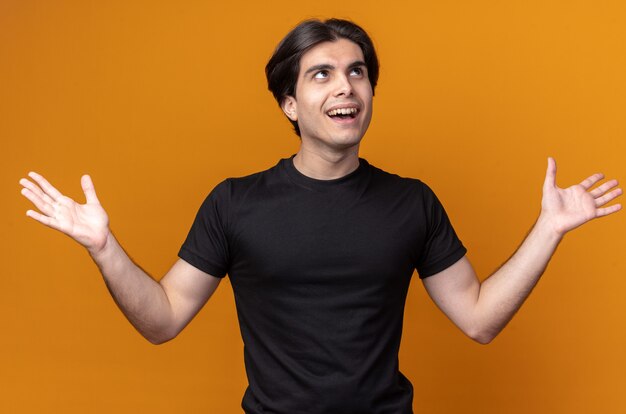 Überraschtes junges hübsches Kerl, das schwarzes T-Shirt trägt, das Hände verteilt auf orange Wand spreizt