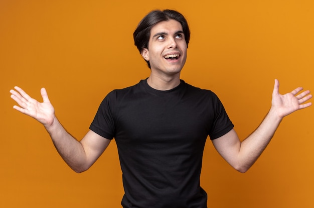 Überraschtes junges hübsches Kerl, das schwarzes T-Shirt trägt, das Hände verteilt auf orange Wand spreizt