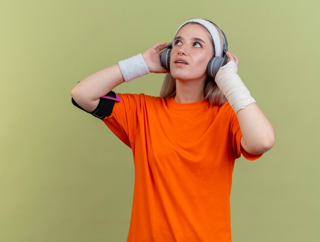 Überraschtes junges kaukasisches sportliches Mädchen mit Hosenträgern auf Kopfhörern, das Stirnband-Armbänder und Telefonarmband trägt, schaut nach oben