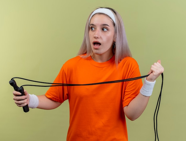 Überraschtes junges kaukasisches sportliches Mädchen mit Hosenträgern, das Stirnband und Armbänder trägt, hält Springseil mit Blick auf die Seite