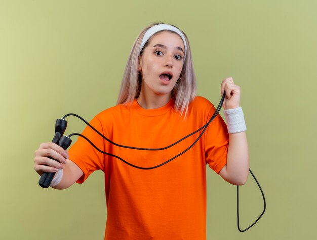 Überraschtes junges kaukasisches sportliches Mädchen mit Hosenträgern, das Stirnband und Armbänder trägt, hält Springseil