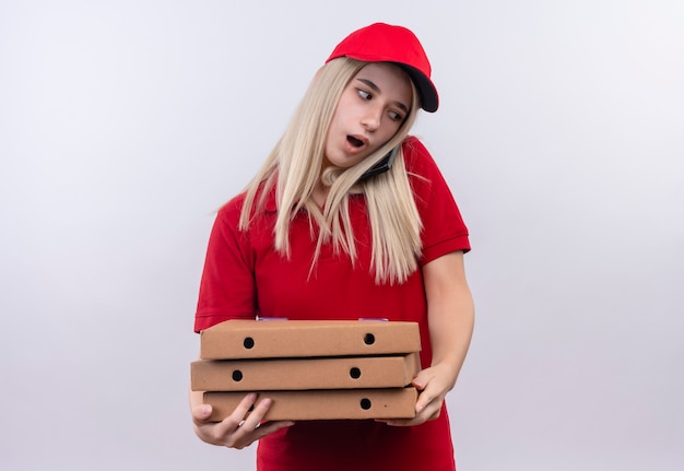 Kostenloses Foto Überraschtes junges mädchen der lieferung, das rotes t-shirt und kappe hält, die pizzakiste hält und am telefon auf lokalem weißem hintergrund spricht