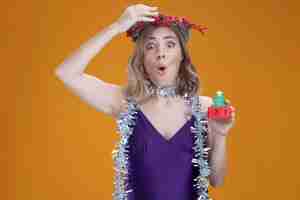 Kostenloses Foto Überraschtes junges schönes mädchen, das lila kleid und kranz mit girlande am hals trägt, das weihnachtsspielzeug hält, das hand auf den kopf einzeln auf braunem hintergrund legt