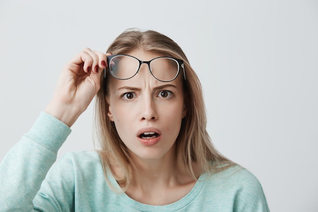 Kostenloses Foto Überraschtes junges weibliches model mit langen blonden haaren, brille und blauem langarmhemd, sieht entsetzt aus