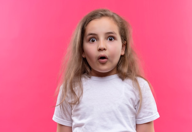 Überraschtes kleines Schulmädchen, das weißes T-Shirt auf lokalisiertem rosa Hintergrund trägt