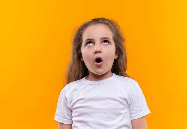 Überraschtes kleines Schulmädchen, das weißes T-Shirt trägt, das oben auf lokalisiertem orange Hintergrund schaut
