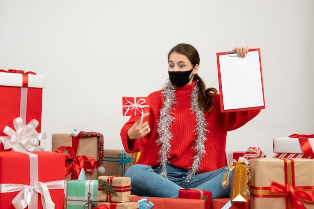 überraschtes Mädchen mit schwarzer Maske, die Dokumente und Geschenk hält, das herum Geschenke auf Weiß sitzt