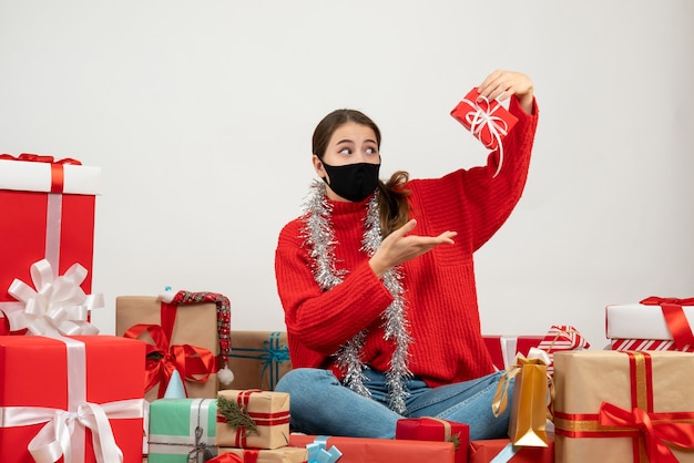 überraschtes Mädchen mit schwarzer Maske, die Geschenk sitzt, das Geschenke auf Weiß sitzt