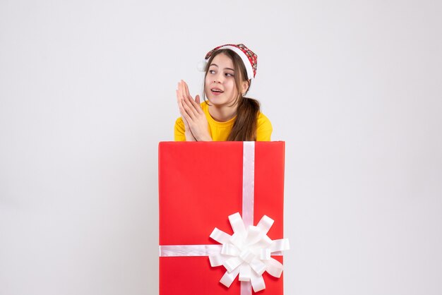 überraschtes Mädchen mit Weihnachtsmütze, die hinter großem Weihnachtsgeschenk auf Weiß steht