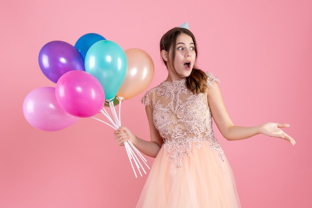 überraschtes Partygirl mit Partykappe, das Luftballons auf rosa hält