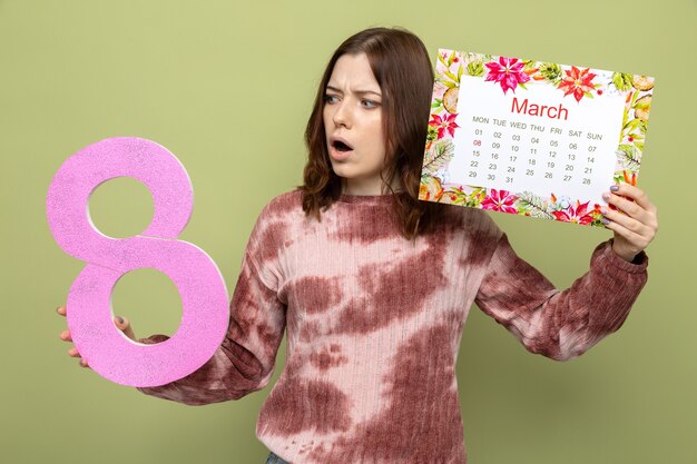 Überraschtes schönes junges mädchen am glücklichen frauentag, der nummer acht mit kalender hält