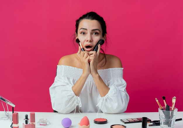 Überraschtes schönes mädchen sitzt am tisch mit make-up-werkzeugen hält und setzt auf gesicht make-up-pinsel isoliert auf rosa wand