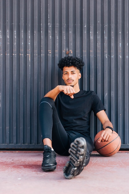 Kostenloses Foto Überzeugter ethnischer junger mann, der auf straße mit basketball sitzt