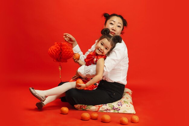 Umarmen, glücklich lächeln, Laternen halten. Frohes chinesisches Neujahr 2020. Asiatisches Mutter- und Tochterporträt auf rotem Hintergrund in traditioneller Kleidung.