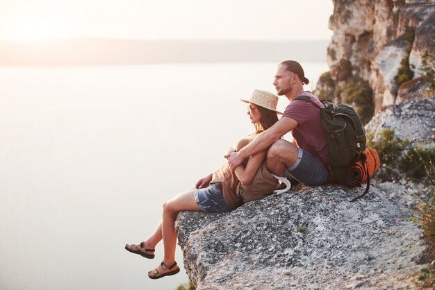 Umarmendes Paar mit Rucksack, der oben auf Felsenberg sitzt und Aussichtsküste einen Fluss oder See genießt.