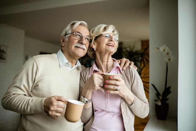 Umarmtes älteres Paar, das durch das Fenster schaut, während es zu Hause Kaffee trinkt