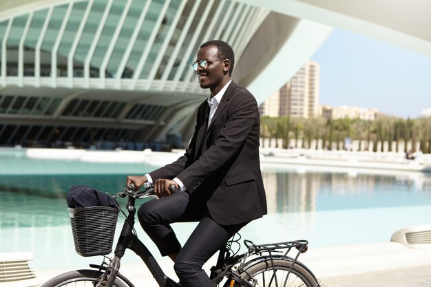 Umweltfreundlicher schwarzer Manager im formellen Anzug und in der Sonnenbrille, die sich freuen, während sie Fahrrad fahren, um in der städtischen Umgebung zu arbeiten, glücklich lächelnd. Geschäft, Lebensstil, Transport und Menschen