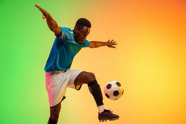 Unaufhaltsamer afroamerikanischer männlicher fußball-fußballspieler, der in aktion isoliert auf gradientenstudiohintergrund im neonlicht trainiert. konzept der bewegungsaktionen für einen gesunden lebensstil