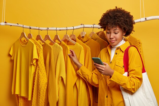 Unbekannte dunkelhäutige Frau mit Afro-Frisur, steht in der Nähe von gelben Kleiderständern, trägt Einkaufstasche auf der Schulter, wählt neues Outfit aus