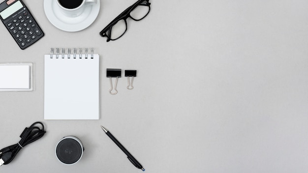 Unbelegter gewundener Notizblock umgeben mit Taschenrechner; Teetasse; Büroklammer; Redner; Stift; Kabel und Brille über grauem Hintergrund