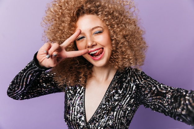 Unbeschwerte Dame mit blauen Lidschatten zeigt Zunge und Zeichen des Friedens. Frau im Disco-Outfit nimmt Selfie auf isoliertem Raum.