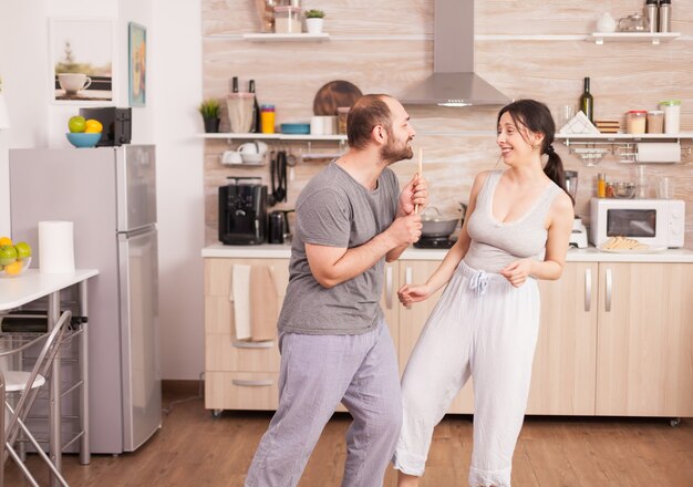 Unbeschwertes, glückliches und fröhliches Paar, das am Morgen in der Küche tanzt und singt. Fröhlicher Ehemann und Ehefrau lachen, singen, tanzen, hören nachdenklich, leben glücklich und sorgenfrei. Positive Menschen