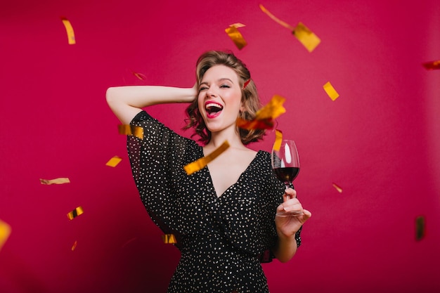 Unbeschwertes junges weibliches Modell in Partykleidung, das Wein unter Glitzerkonfetti trinkt. Lachende weiße Frau mit kurzem Haarschnitt, die bei der Veranstaltung tanzt.