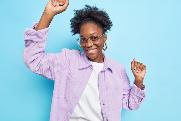 Unbeschwertes positives Teenager-Mädchen mit dunkler Haut, Afro-Haar und zahnigem Lächeln trägt transparente Brille, Freizeithemd isoliert auf Blau