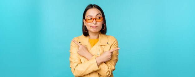 Unentschlossene asiatische Frau mit Sonnenbrille trifft die Wahl zwischen zwei Varianten, zeigt seitlich und sieht verwirrt über blauem Hintergrund aus