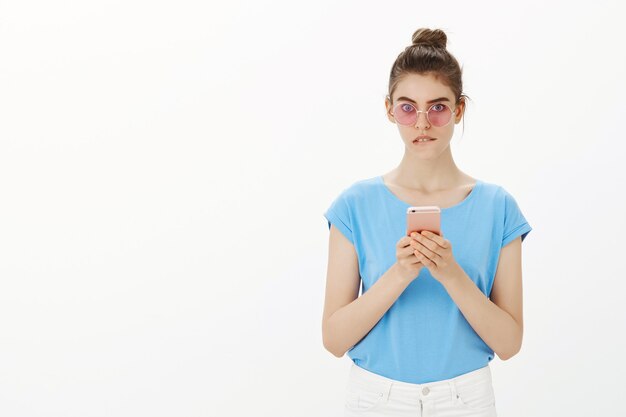 Unentschlossene Frau in der Sonnenbrille beißt sich auf die Lippe, während sie das Mobiltelefon benutzt