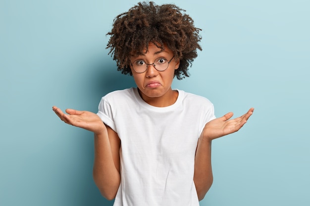 Unentschlossenes dunkelhäutiges Mädchen breitet Palmen mit verwirrtem Ausdruck aus, trägt lässiges Mock-up-T-Shirt, runzelt die Stirn angesichts der Unsicherheit, hat Afro-Haarschnitt, isoliert über blauer Wand. Frau steht vor Schwierigkeiten