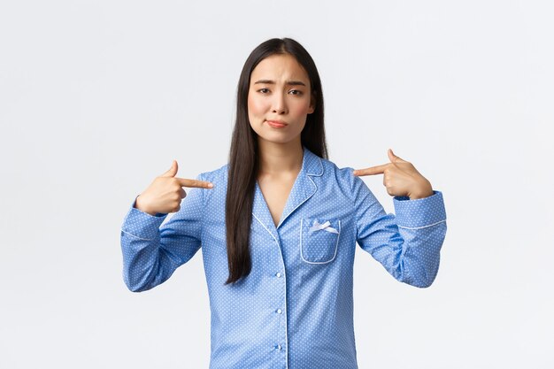 Unentschlossenes und unsicheres asiatisches Mädchen im blauen Pyjama grinst unsicher und zeigt auf sich selbst, ist sich seiner eigenen Fähigkeiten nicht sicher, spricht über sich selbst, steht auf weißem Hintergrund