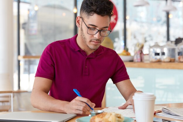 Unerfahrener junger männlicher Arbeiter erledigt Fernarbeit, hält blauen Stift, schreibt Aufzeichnungen oder Notizen in Notizblock, macht Planungen für nächste Woche. Student bereitet sich auf die College-Prüfung vor, sitzt im Restaurant