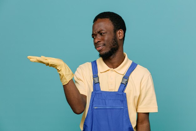 Unerfreut, so zu tun, als würde er etwas junges afroamerikanisches Putzermännchen in Uniform mit Handschuhen isoliert auf blauem Hintergrund halten