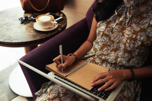 Unerkennbare Frau, die im Café mit Laptop sitzt und in Notizbuch schreibt