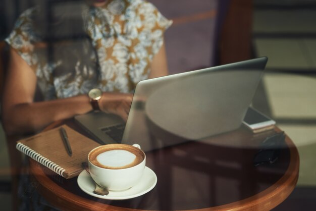 Unerkennbare junge Frau, die bei Tisch im Café sitzt und an Laptop arbeitet