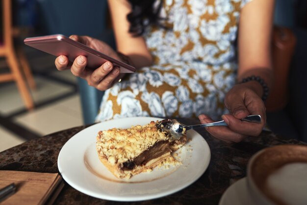 Unerkennbare junge Frau, die im Café sitzt, Smartphone verwendet und Streuseltorte isst