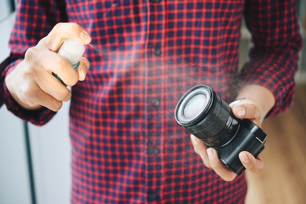 Kostenloses Foto unerkennbarer männlicher fotograf, der kameraobjektiv mit reinigungsflüssigkeit sprüht