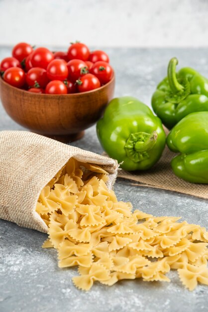 Ungekochte Nudeln in einer rustikalen Tasche mit Kirschtomaten und Paprika auf Marmortisch.