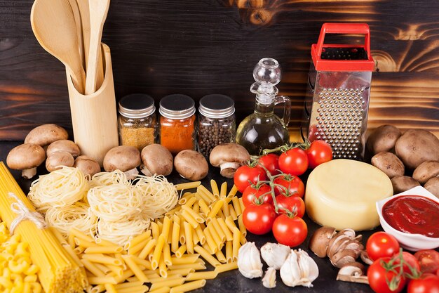 Ungekochte rohe Makkaroni, Nudeln und Spaghetti neben frischem und gesundem Gemüse, verschiedenen Gewürzen und Sonnenblumenöl auf dunklem rustikalem Holzhintergrund