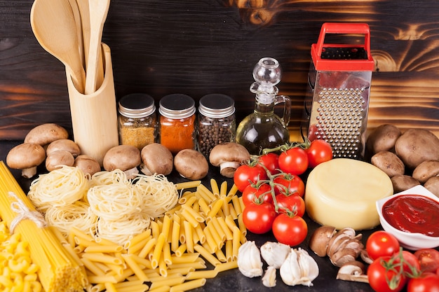 Ungekochte rohe Makkaroni, Nudeln und Spaghetti neben frischem und gesundem Gemüse, verschiedenen Gewürzen und Sonnenblumenöl auf dunklem rustikalem Holzhintergrund