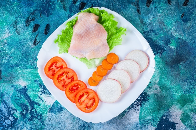 Ungekochtes Hühnerfleisch neben Tomaten, Rettich und Karotten auf einem Teller