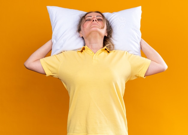 Ungesunde Frau in gelbem Hemd, die an Grippe und Erkältung leidet und sich mit Kissen unter dem Kopf unwohl fühlt, schläft über orangefarbener Wand