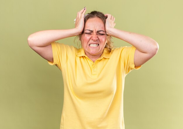 Ungesunde Frau in gelbem Hemd, die sich schrecklich fühlt, wenn sie ihren Kopf berührt und unter starken Kopfschmerzen leidet, die eine Grippe über der grünen Wand haben?