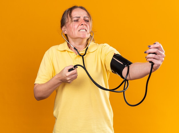 Ungesunde Frau in gelbem Hemd, die sich unwohl fühlt und an Grippe und Erkältung leidet, die ihren Blutdruck mit einem Tonometer misst und besorgt über der orangefarbenen Wand steht