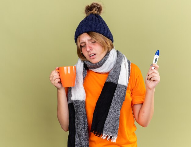 Ungesunde junge Frau, die Hut mit Schal um den Hals trägt und sich unwohl fühlt, wenn sie heißen Tee trinkt und ein digitales Thermometer hält, das an einer Grippe leidet, die über der grünen Wand steht?