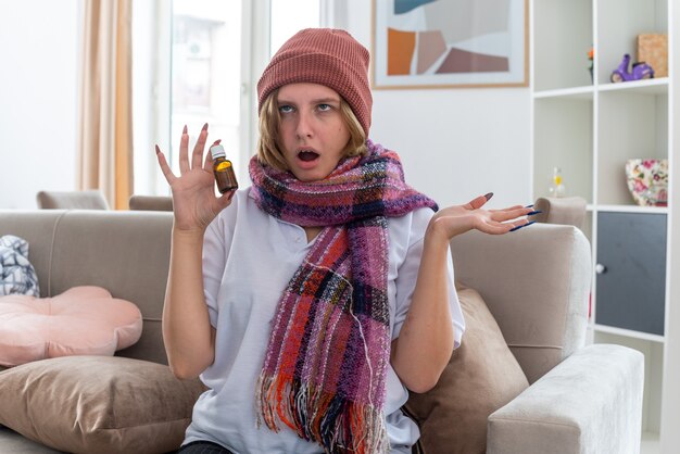 Ungesunde junge Frau im Hut mit warmem Schal um den Hals, der sich unwohl und krank fühlt und an Erkältung und Grippe leidet, die gestresste und genervte Medizinflasche hält, die auf Couch in hellem Wohnzimmer sitzt