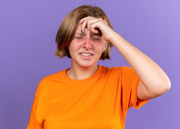 Ungesunde junge Frau in orangefarbenem T-Shirt, die sich schrecklich fühlt, wenn sie ihren Kopf berührt, während sie sich schwindelig fühlt und Grippe über lila Wand steht?