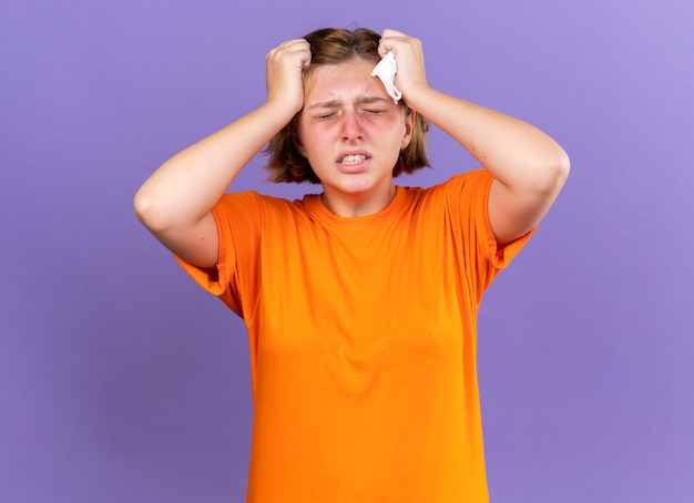 Ungesunde junge Frau in orangefarbenem T-Shirt, die sich schrecklich fühlt, wenn sie ihren Kopf berührt, während sie sich schwindelig fühlt und unter starken Kopfschmerzen leidet